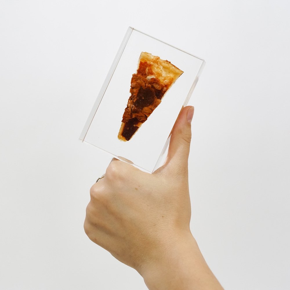 A hand holding a block of acrylic with a slice of pizza embedded inside.