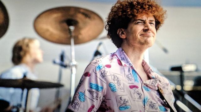 Joseph Mazzello as John Deacon, with a very 80's pink printed shirt and big curly hair.