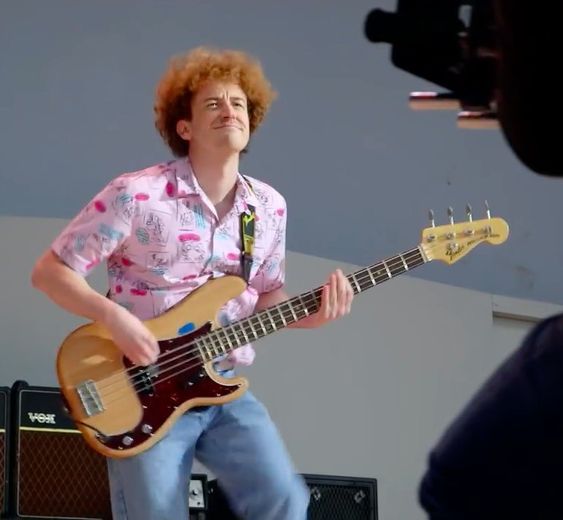 Joseph Mazzello as John Deacon, with a very 80's pink printed shirt and big curly hair, rocking out on the bass.