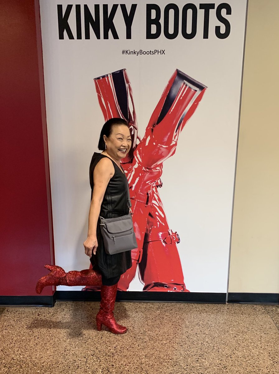 Small Asian lady in tall sparkly red boots in front of a Kinky Boots poster.