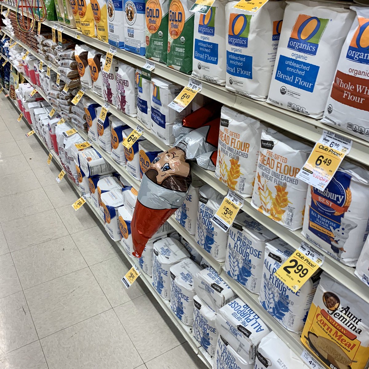 A deflating elf on the shelf mylar balloon pokes his head out of a grocery store shelf of flour.