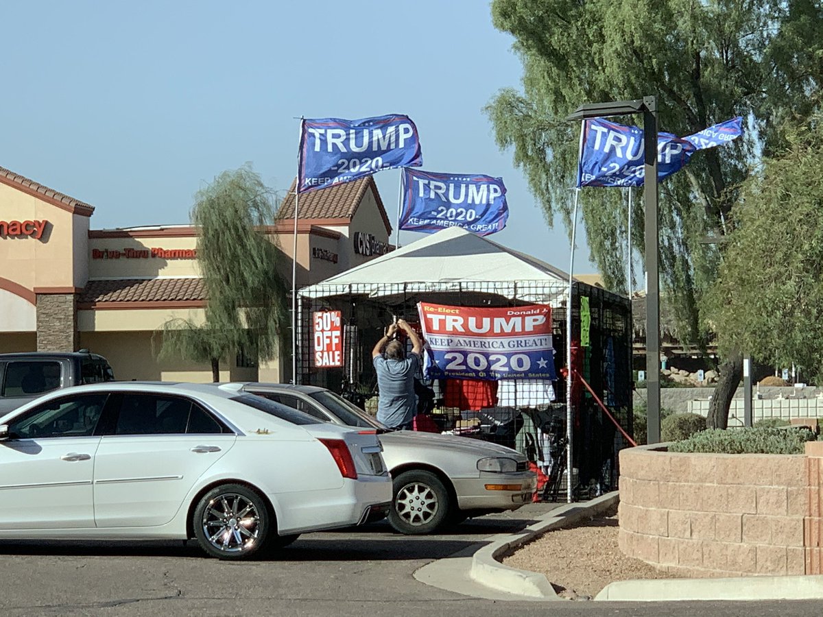 Man setting up Trump merch tent with sad 50% off sale sign.