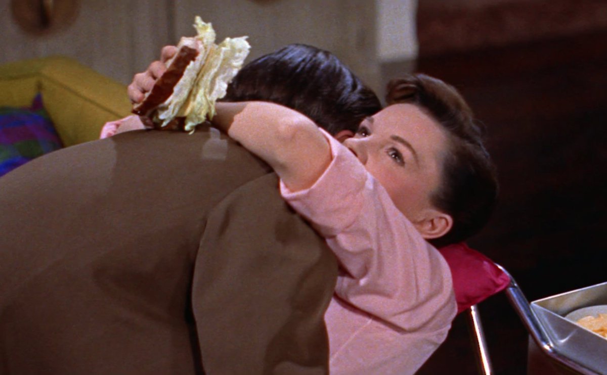 Judy Garland is embracing a man but she looks distracted and has a giant, lettuce-filled sandwich in her hand