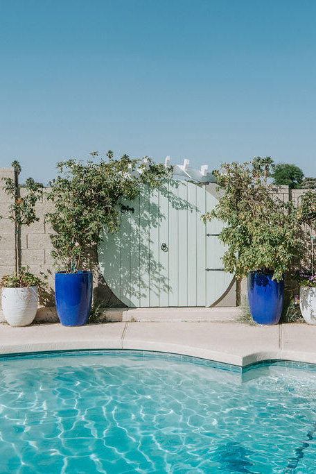 a backyard with sparkling pool, lots of plants, and on the cement block wall, a gate has been built to look like a round hobbit door