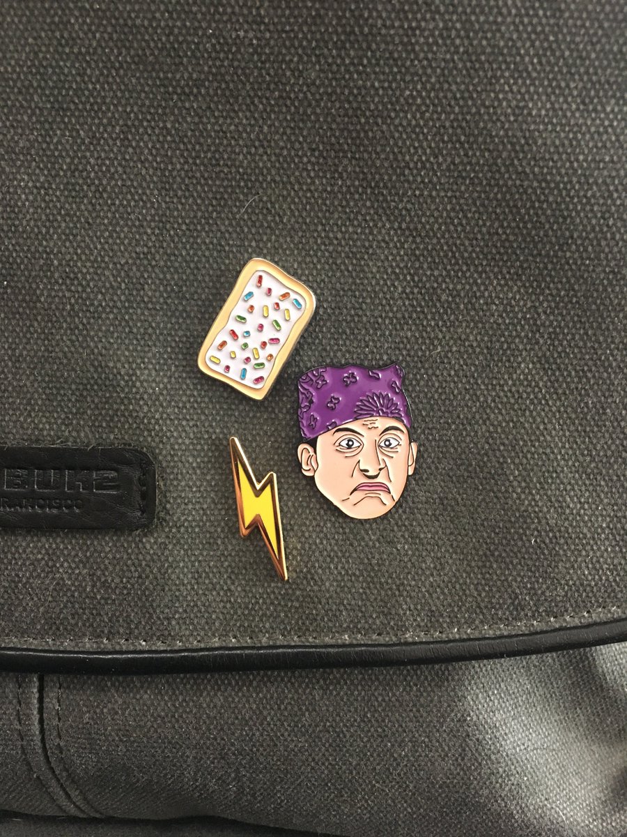 Closeup shot of a backpack with three enamel pins: a strawberry Pop Tart, a gold lightning bolt, and the Michael Scott character "Prison Mike" from the Office.