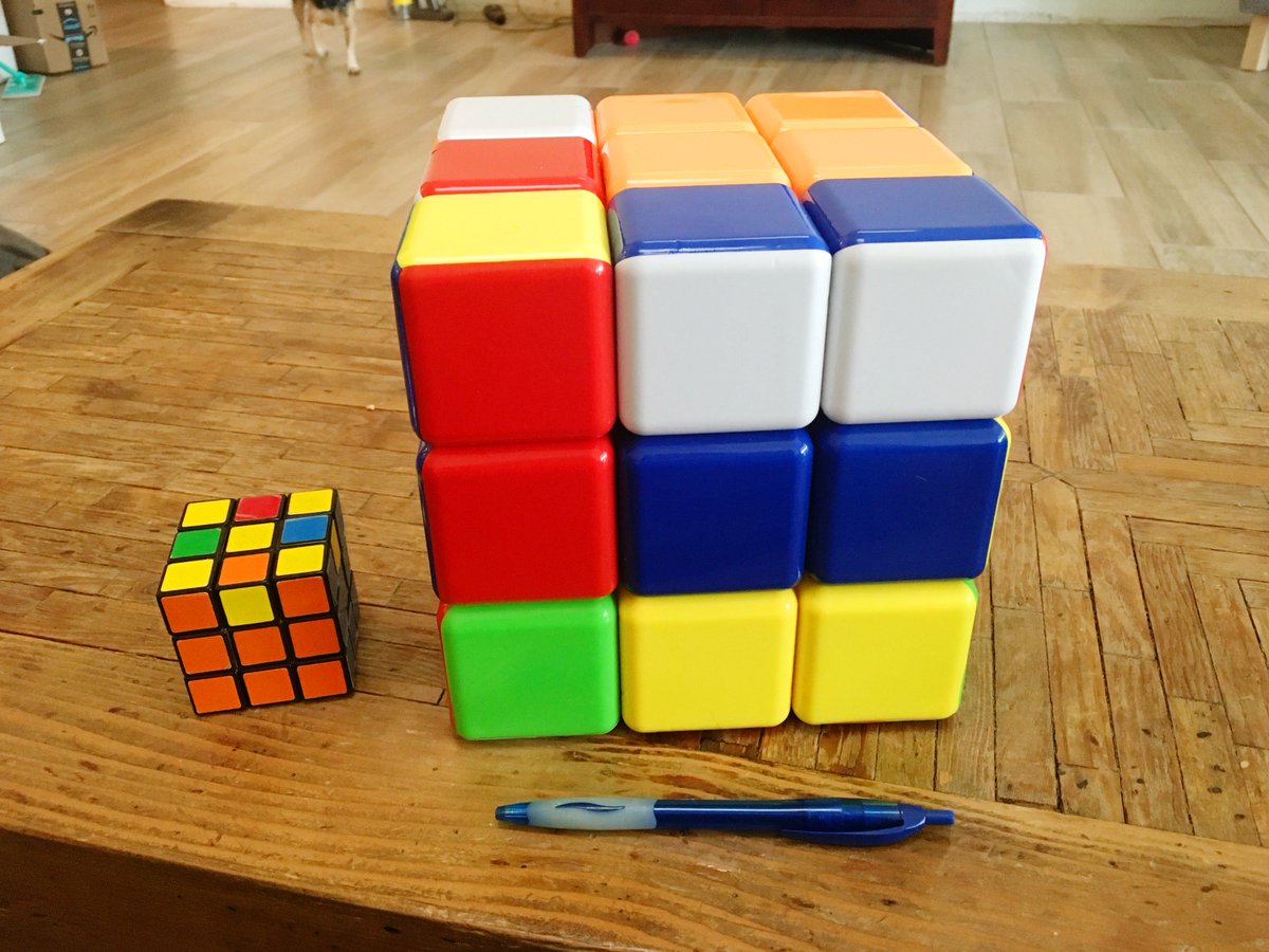 A giant Rubik's cube next to an original Rubik's cube and a pen. Each individual piece of the giant cube is the size of the entire original cube.